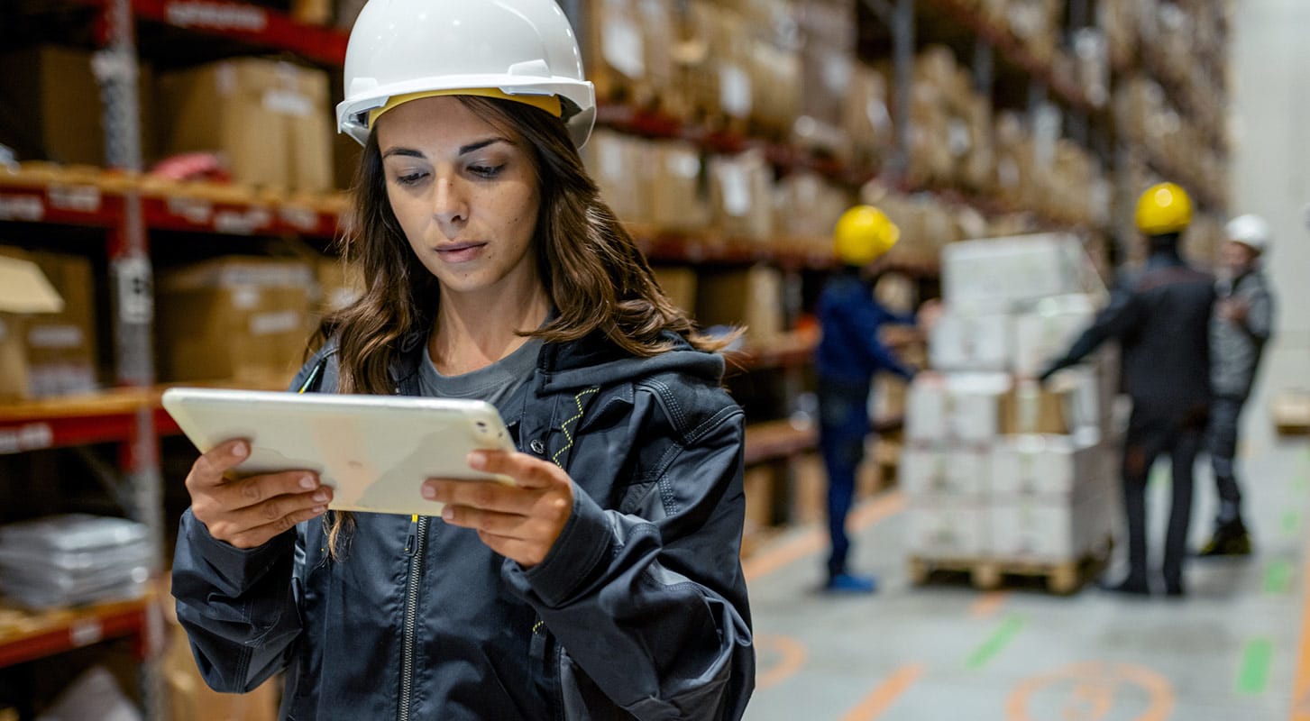 A female worker in a hard hat and dark jacket looks at a tablet in a warehouse, with shelves of boxes and other workers in the background.